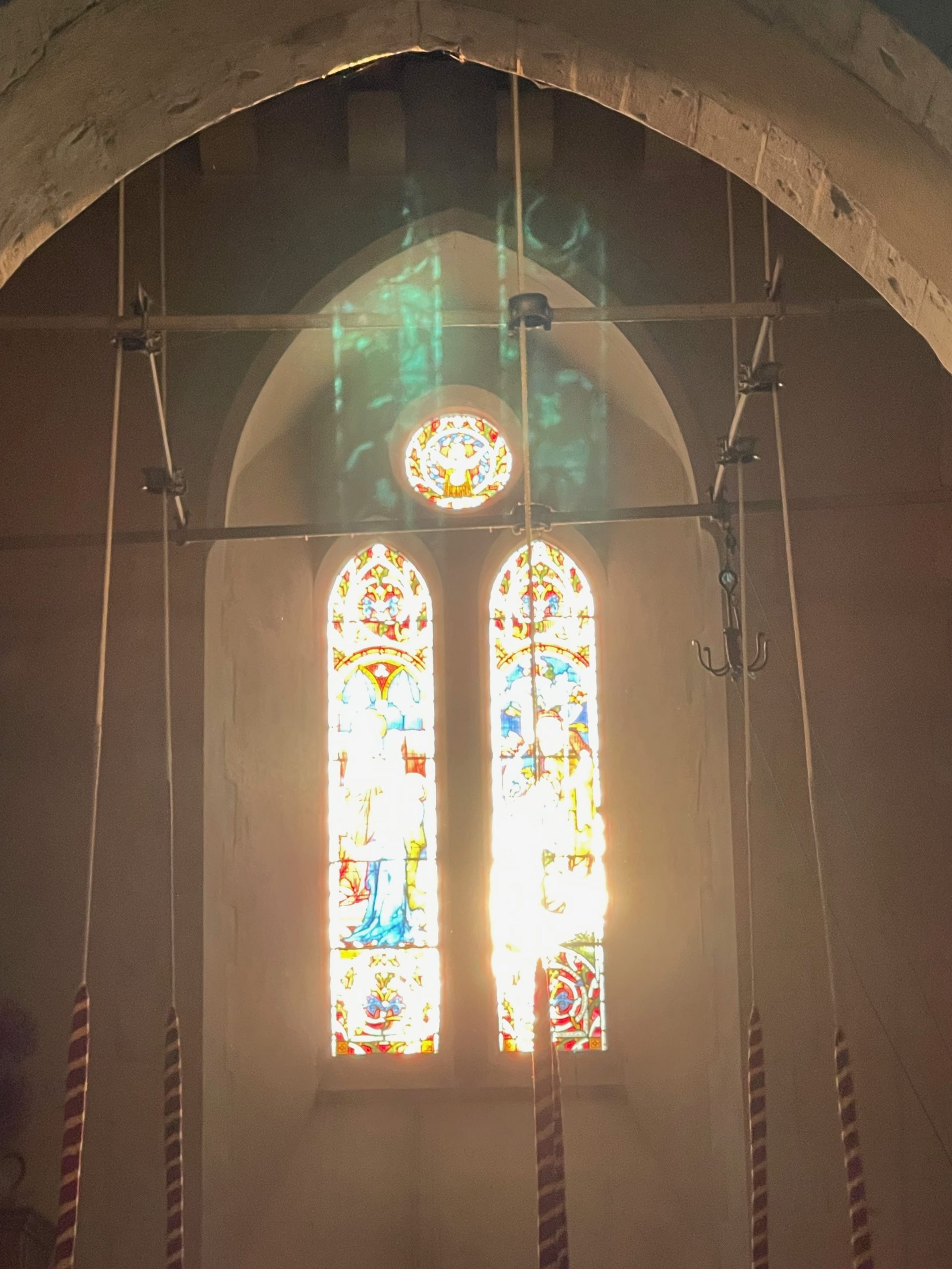 Bell-ringing ropes hang in front of a stained-glass church window illuminated by bright sunlight. Green reflections from the stained glass are cast onto the stone arch and wall above.