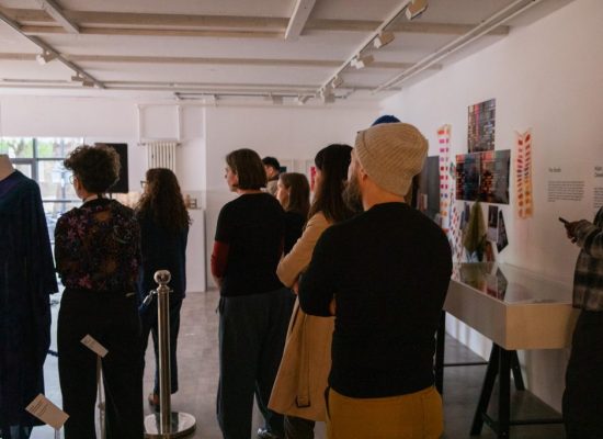 Visitors view exhibits in a gallery space, including a garment displayed on a mannequin and exhibition panels mounted on the walls. The room is brightly lit, with several people standing and observing the displays.