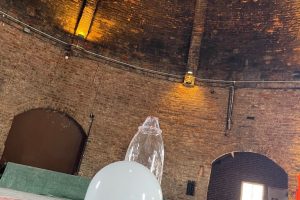 White glass forms and test pieces are arranged on the floor inside a circular brick kiln. The kiln's curved walls and high dome create an industrial studio setting for glassmaking experiments.