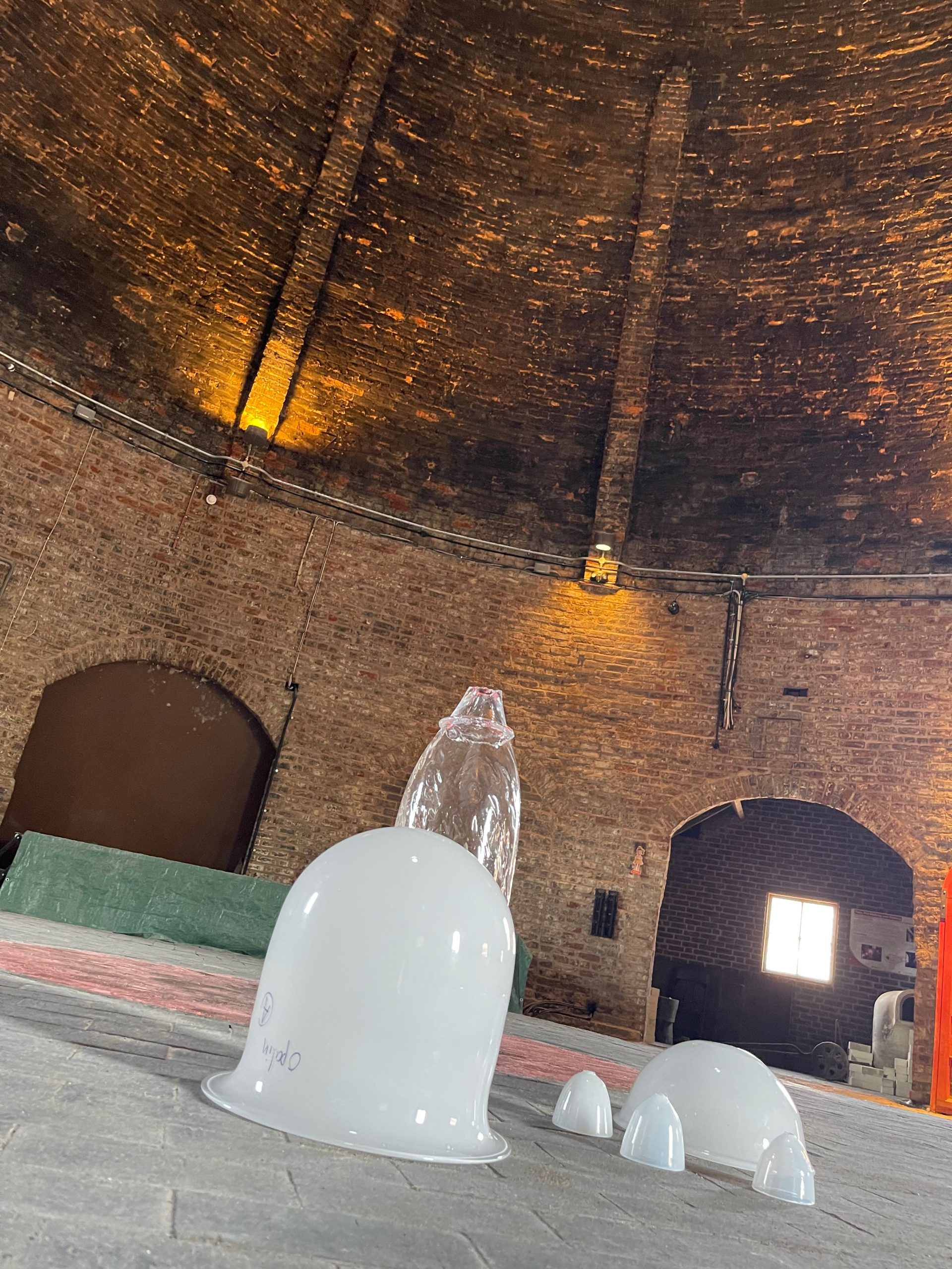 White glass forms and test pieces are arranged on the floor inside a circular brick kiln. The kiln's curved walls and high dome create an industrial studio setting for glassmaking experiments.