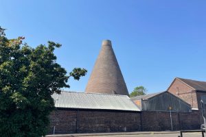 A tall brick bottle kiln rises above industrial buildings beneath a clear blue sky. The historic kiln is viewed from across a street with trees framing the scene.
