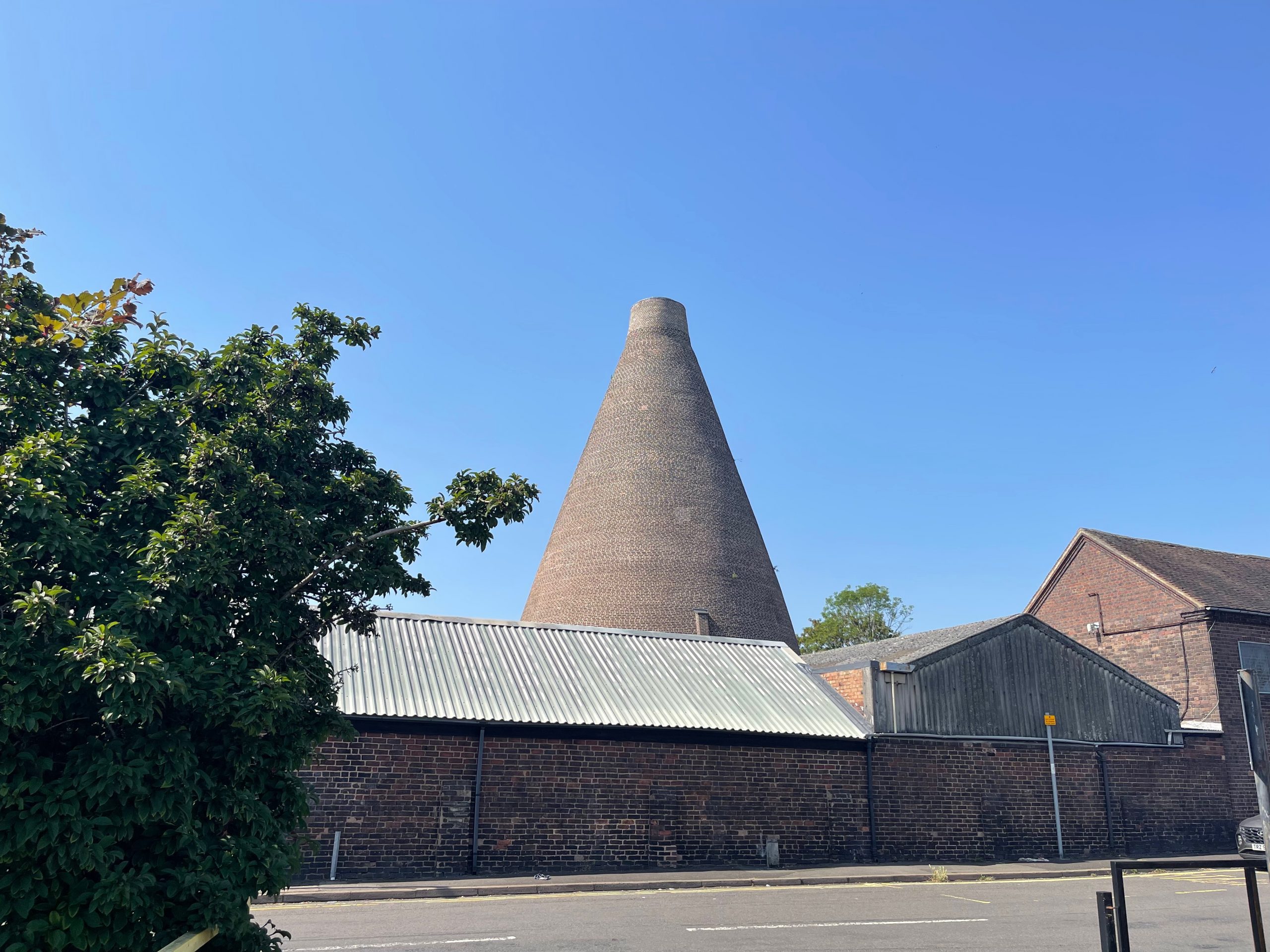 A tall brick bottle kiln rises above industrial buildings beneath a clear blue sky. The historic kiln is viewed from across a street with trees framing the scene.