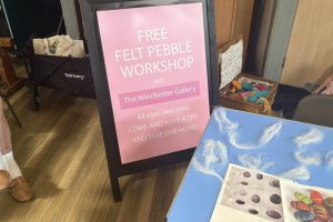 A sign advertises a free felt pebble workshop by The Winchester Gallery, welcoming participants of all ages. Wool fibres and example felted pebble designs are displayed on a nearby table.