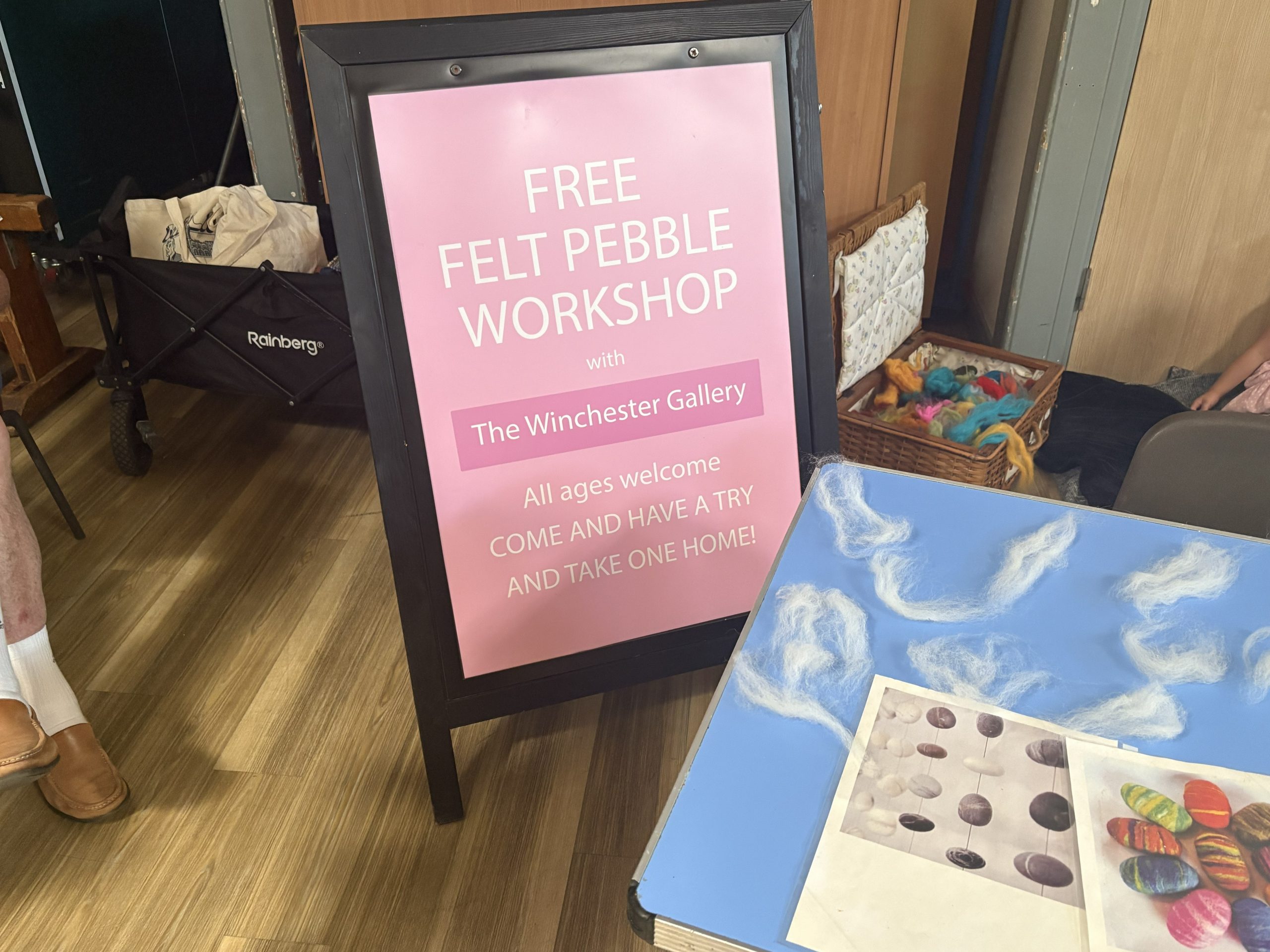 A sign advertises a free felt pebble workshop by The Winchester Gallery, welcoming participants of all ages. Wool fibres and example felted pebble designs are displayed on a nearby table.