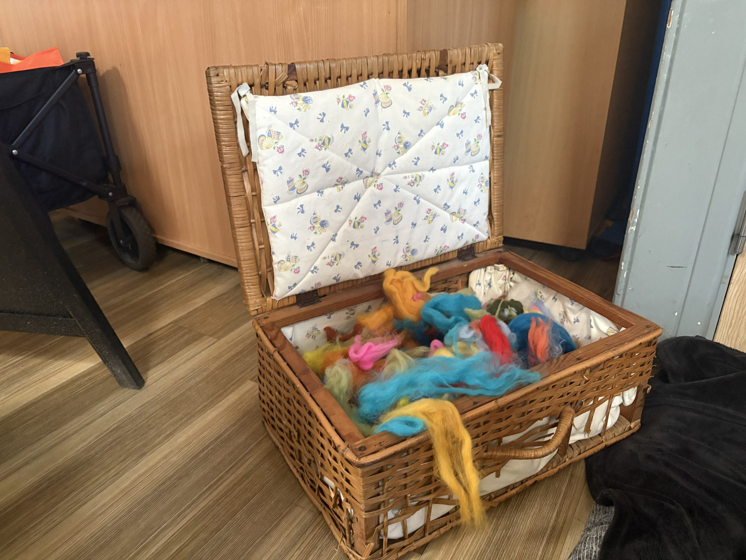An open wicker basket contains colourful wool fibres used for a felting workshop. The materials are arranged indoors ready for participants to use.
