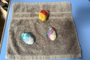 Three handmade felt pebbles in different colour combinations are displayed on a grey towel. The felted shapes show the results of the workshop activity.
