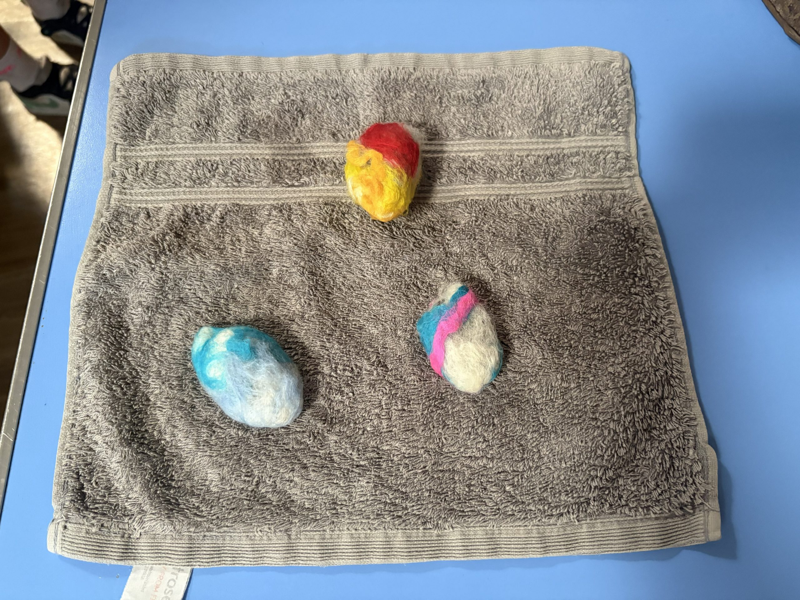 Three handmade felt pebbles in different colour combinations are displayed on a grey towel. The felted shapes show the results of the workshop activity.
