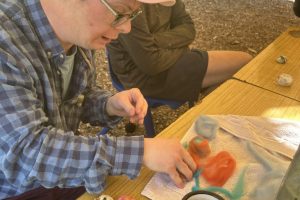 A person wearing a pink cap shapes colorful wool fibers into felted stones at an outdoor table. Another person watches from across the table, with a mug, towel, and crafting materials nearby.