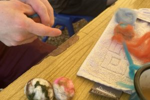 A close-up shot shows hands crafting colourful wool, with completed felted balls resting on a wooden table. Nearby, a towel, colourful fibres, and a blue mug with a tea bag tag are visible in the outdoor setting.