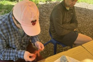 Two individuals sit at a wooden table outdoors, surrounded by green foliage, while engaging in a felt-making craft. One person wears a pink cap and works with orange fibre, while several completed colourful felted balls sit on the table nearby.