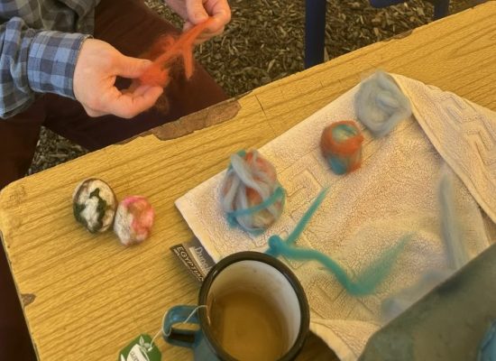 A tabletop felting project with colourful wool roving, two finished felted balls, and a towel beside a mug of tea and a teapot. A person’s hands are stretching orange wool fibre as part of the crafting process.