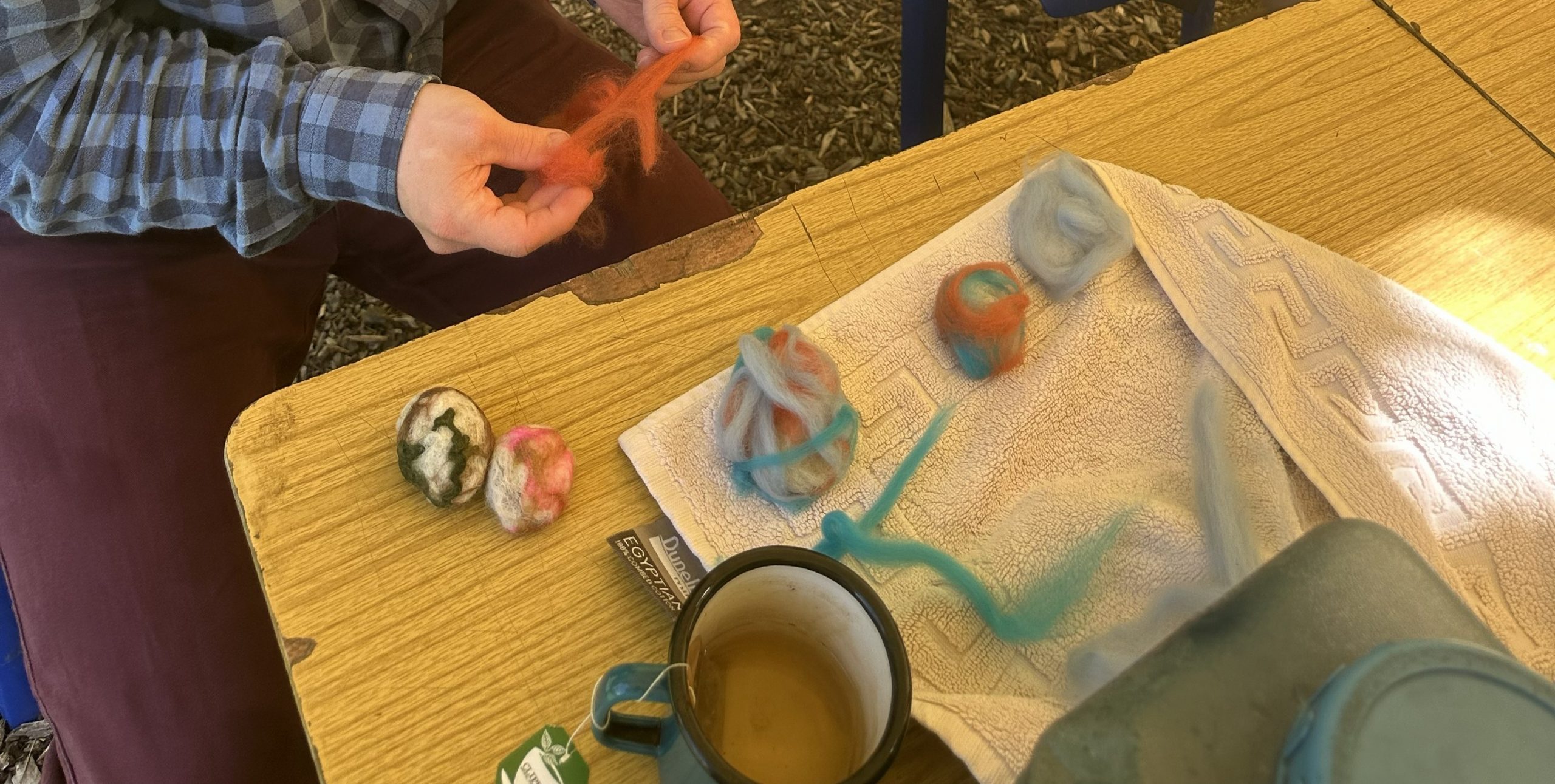 A tabletop felting project with colourful wool roving, two finished felted balls, and a towel beside a mug of tea and a teapot. A person’s hands are stretching orange wool fibre as part of the crafting process.