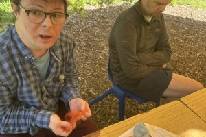 A person wearing a pink cap and glasses looks toward the camera while working with orange fiber at an outdoor crafting table. Beside them, another individual sits with closed eyes, and several colorful felted items rest on the table.