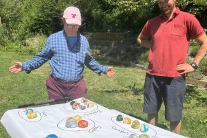 Two people stand outdoors behind a table displaying colourful, handmade felted balls organized into labelled groups on a large sheet of paper. The individual on the left gestures proudly toward the finished projects, while the person on the right watches with a smile.