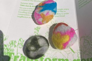 Three finished felted wool shapes in different colour combinations are displayed on an open leaflet. The handmade pieces feature swirls and patterns created from layered wool fibres.