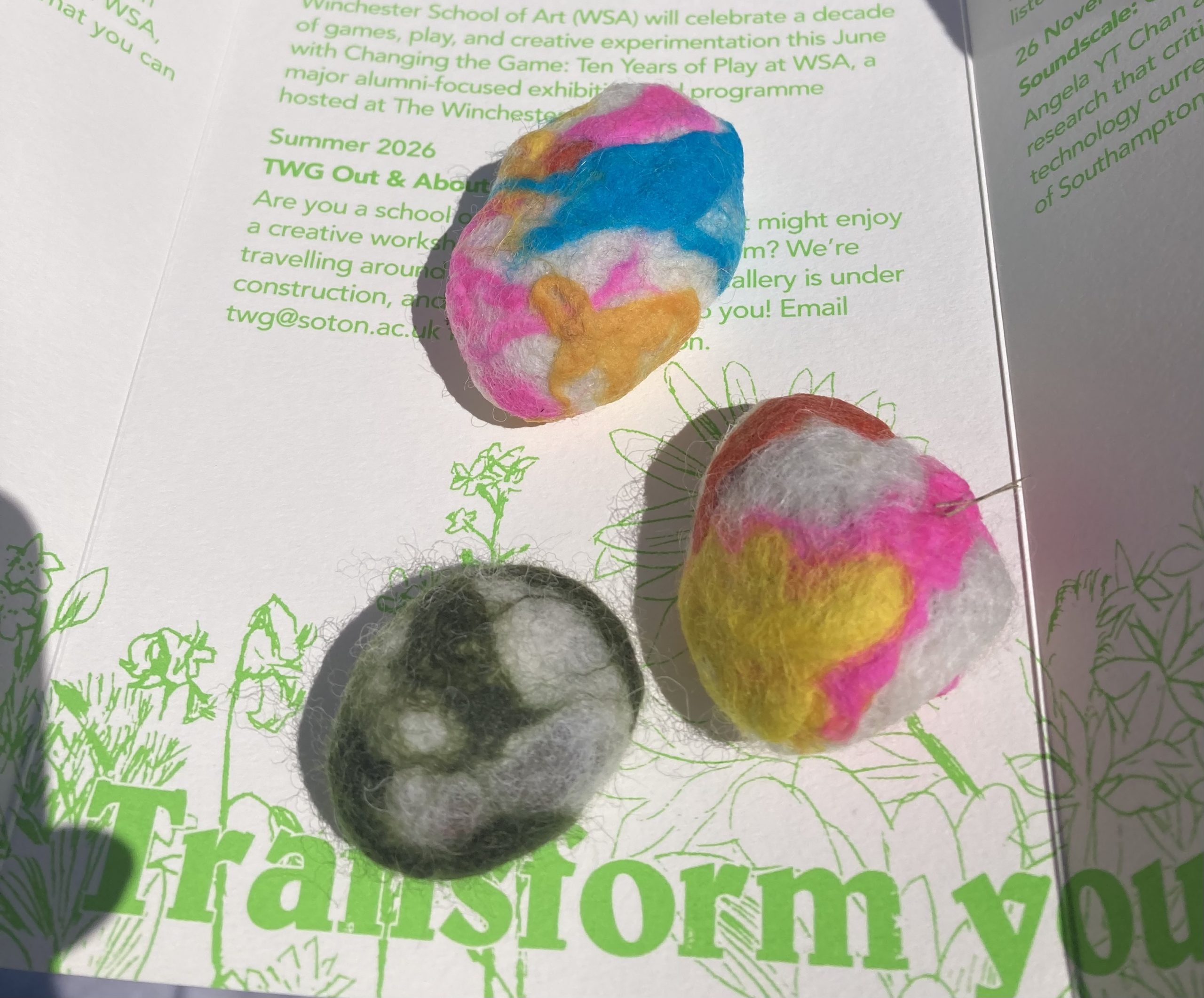 Three finished felted wool shapes in different colour combinations are displayed on an open leaflet. The handmade pieces feature swirls and patterns created from layered wool fibres.