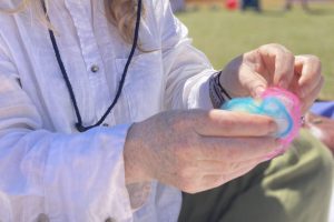 A person demonstrates a felting activity outdoors, holding layers of colourful wool fibres in their hands. The image shows pink, blue, white and yellow fibres being arranged before felting.