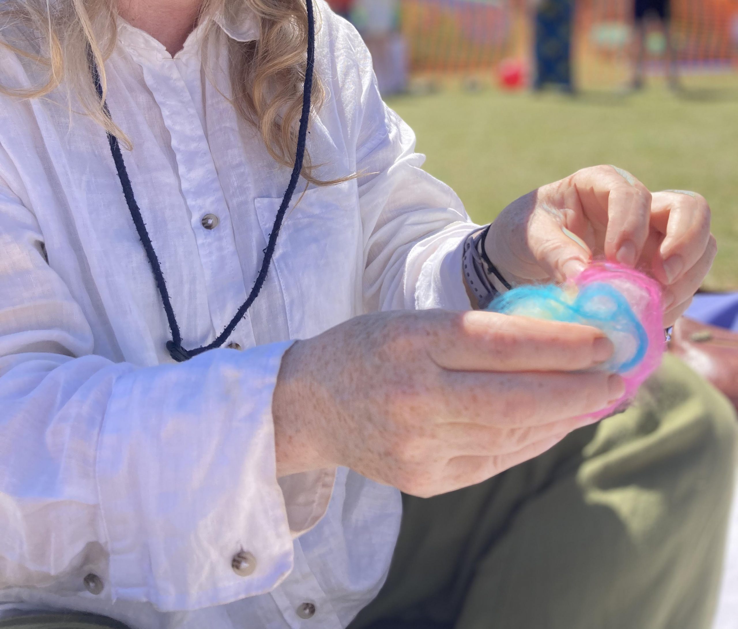 A person demonstrates a felting activity outdoors, holding layers of colourful wool fibres in their hands. The image shows pink, blue, white and yellow fibres being arranged before felting.