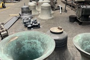 Large bronze bells of varying sizes are arranged across the workshop floor of a bell foundry. Industrial machinery and casting equipment surround the bells inside the spacious factory.