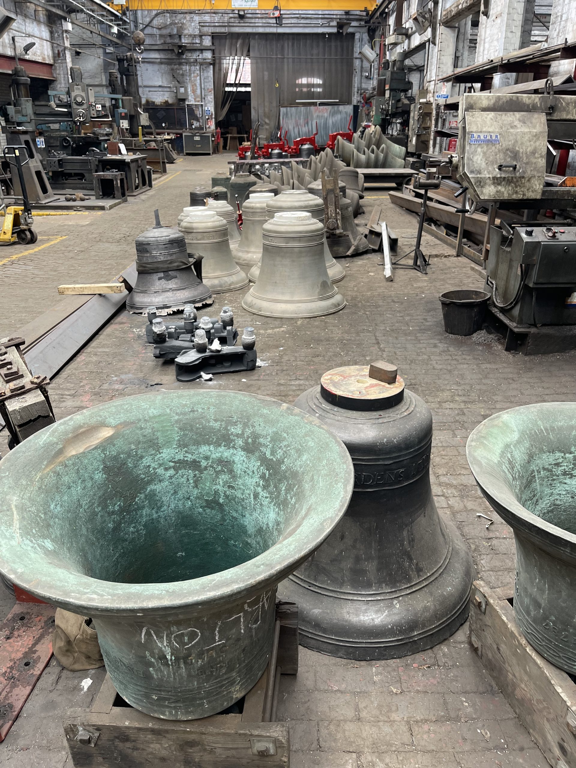Large bronze bells of varying sizes are arranged across the workshop floor of a bell foundry. Industrial machinery and casting equipment surround the bells inside the spacious factory.