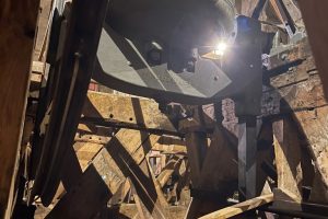 Large church bell mounted within a timber bell frame inside a bell tower. The bell is illuminated by a light shining through the wooden structure.
