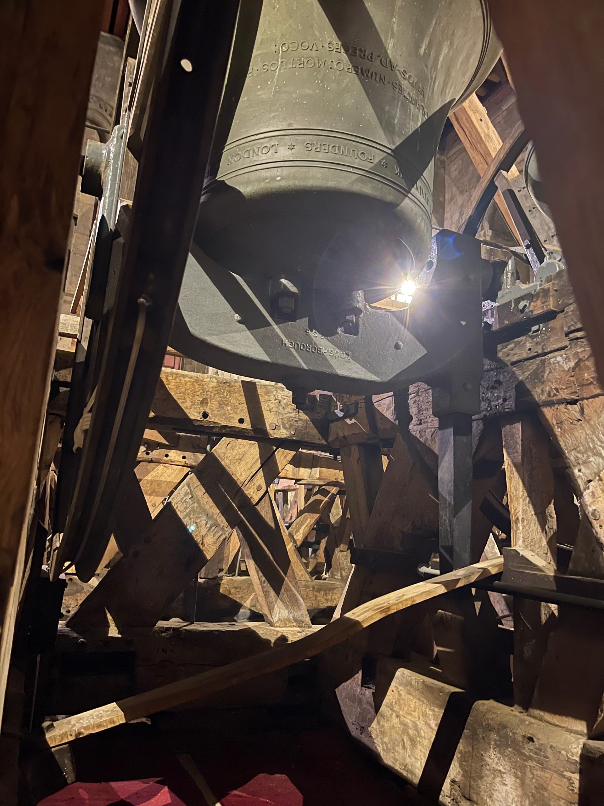 Large church bell mounted within a timber bell frame inside a bell tower. The bell is illuminated by a light shining through the wooden structure.