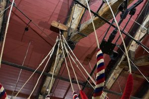 Bell-ringing ropes with red, white and blue striped sallies hang from timber beams in a ringing chamber. The ropes are suspended beneath a wooden ceiling crossed by metal supports.