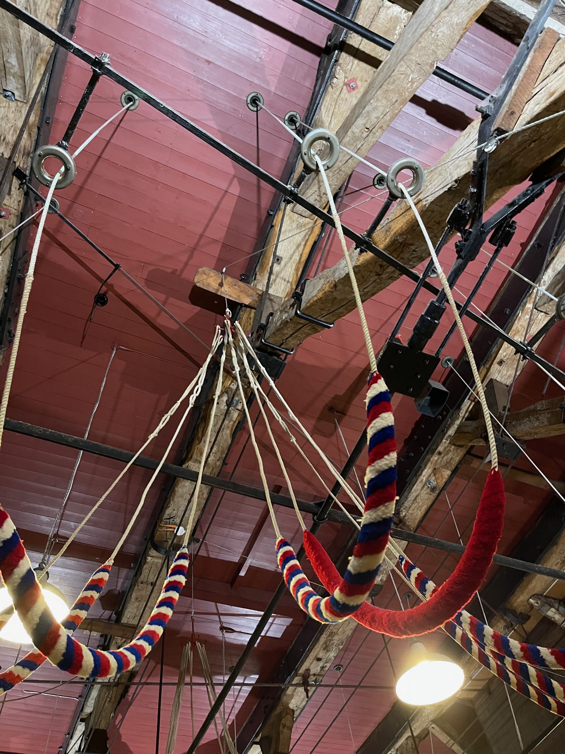 Bell-ringing ropes with red, white and blue striped sallies hang from timber beams in a ringing chamber. The ropes are suspended beneath a wooden ceiling crossed by metal supports.