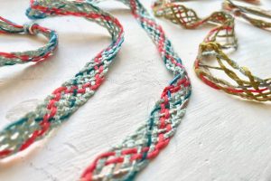 Close-up of braided textile samples in pink, turquoise and cream threads laid across a white surface. The woven cords create delicate looping patterns and textures.