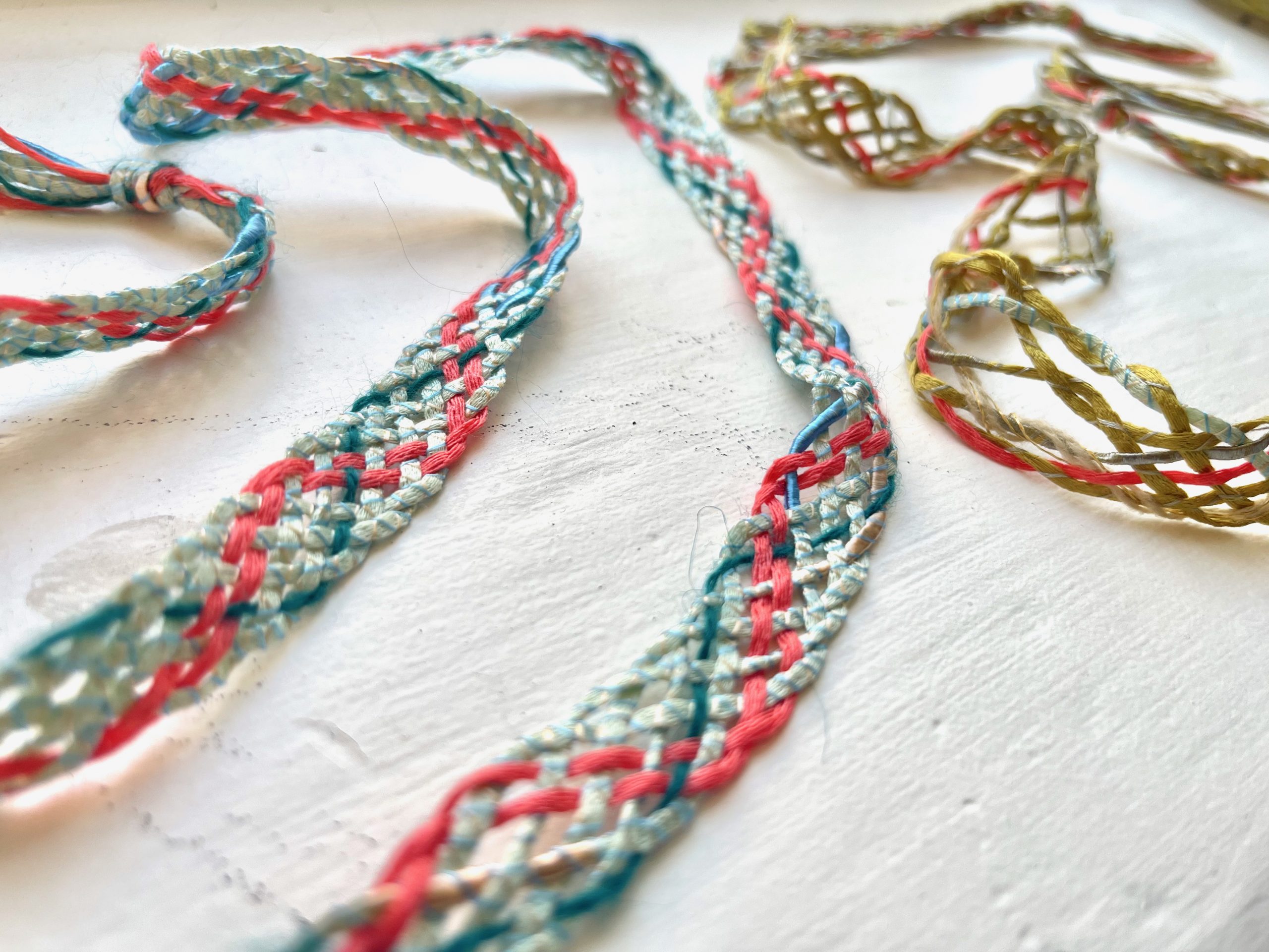 Close-up of braided textile samples in pink, turquoise and cream threads laid across a white surface. The woven cords create delicate looping patterns and textures.