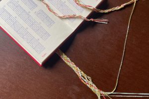 An open bell-ringing methods book lies on a wooden table alongside colourful braided cords woven through and around its pages. The braids echo the structured patterns printed in the book.