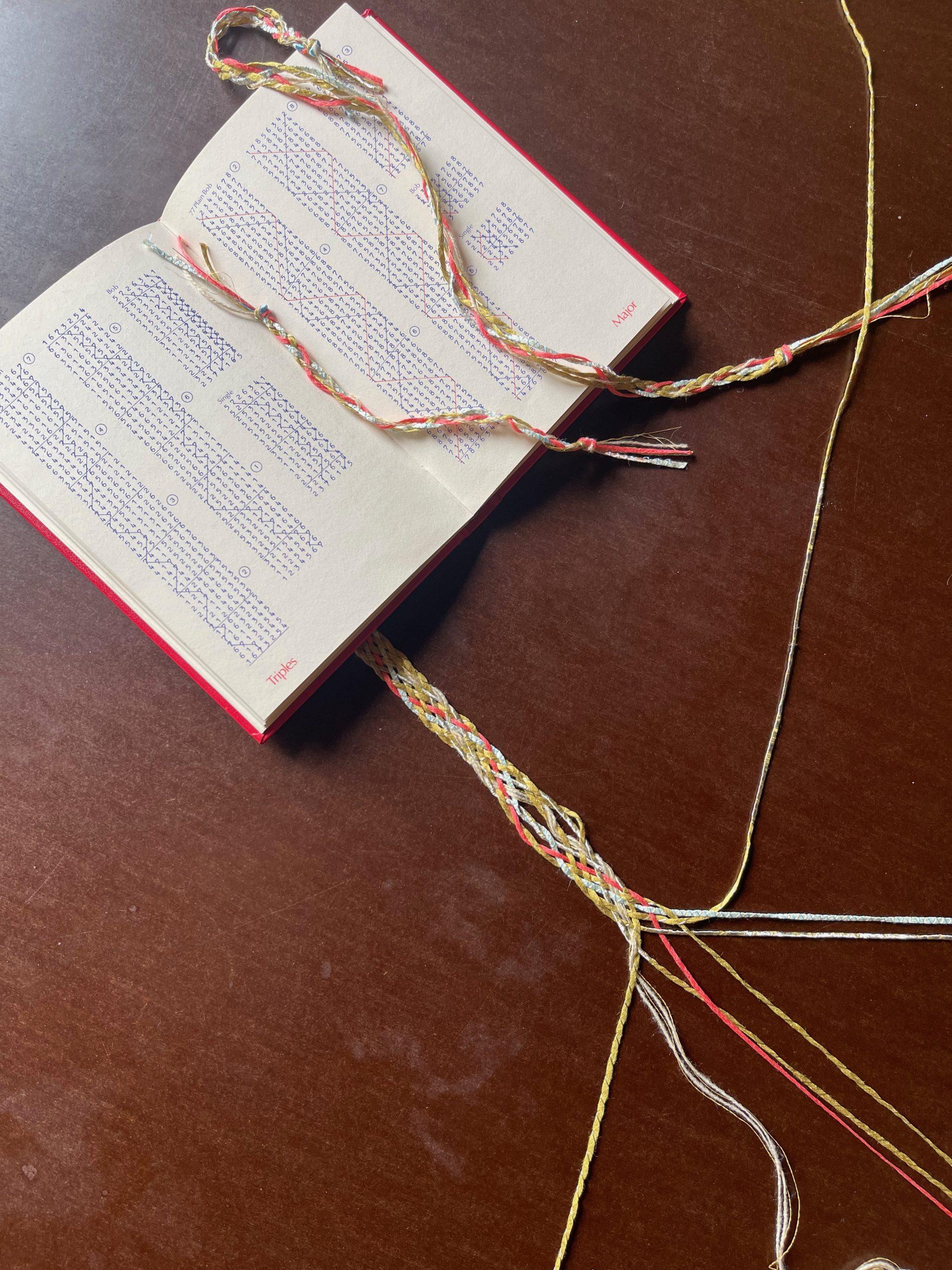 An open bell-ringing methods book lies on a wooden table alongside colourful braided cords woven through and around its pages. The braids echo the structured patterns printed in the book.