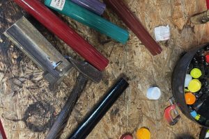 Colourful glass rods and glassblowing tools are arranged across a workshop workbench. Samples of red, yellow, blue, turquoise and white glass are displayed alongside equipment used for shaping glass.