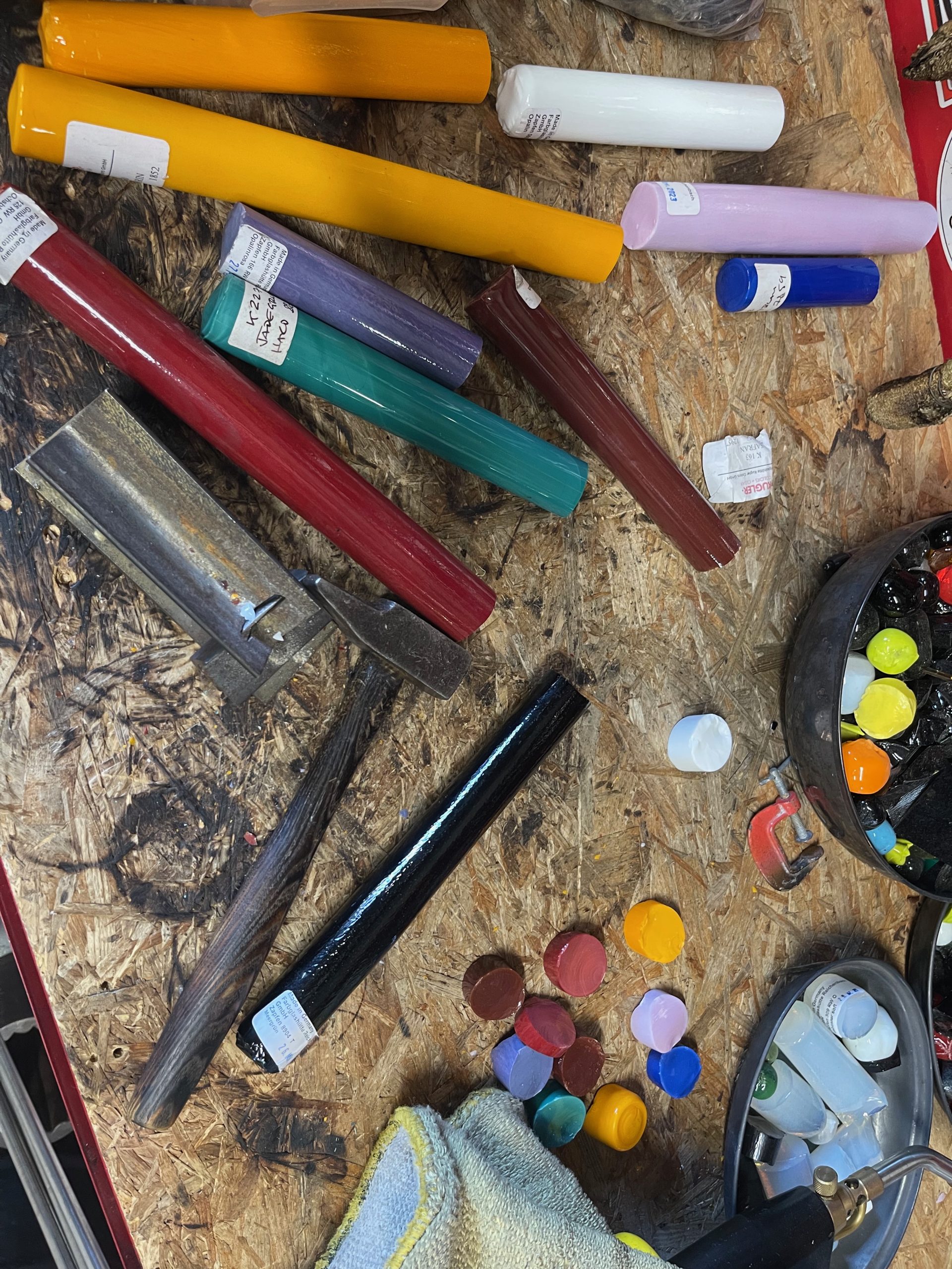Colourful glass rods and glassblowing tools are arranged across a workshop workbench. Samples of red, yellow, blue, turquoise and white glass are displayed alongside equipment used for shaping glass.