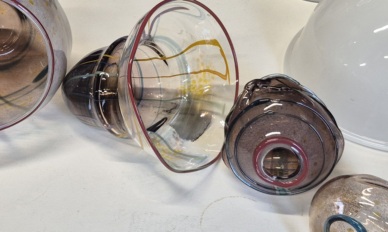 Close-up of hand-blown glass forms with translucent surfaces and coloured lines, arranged on a worktable. The vessel shapes resemble bell-like bubbles and experimental glass studies.