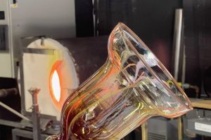 Close-up of a hand-blown glass vessel being shaped on a blowpipe in a glassmaking studio. The transparent form is illuminated by the glow of a furnace in the background.