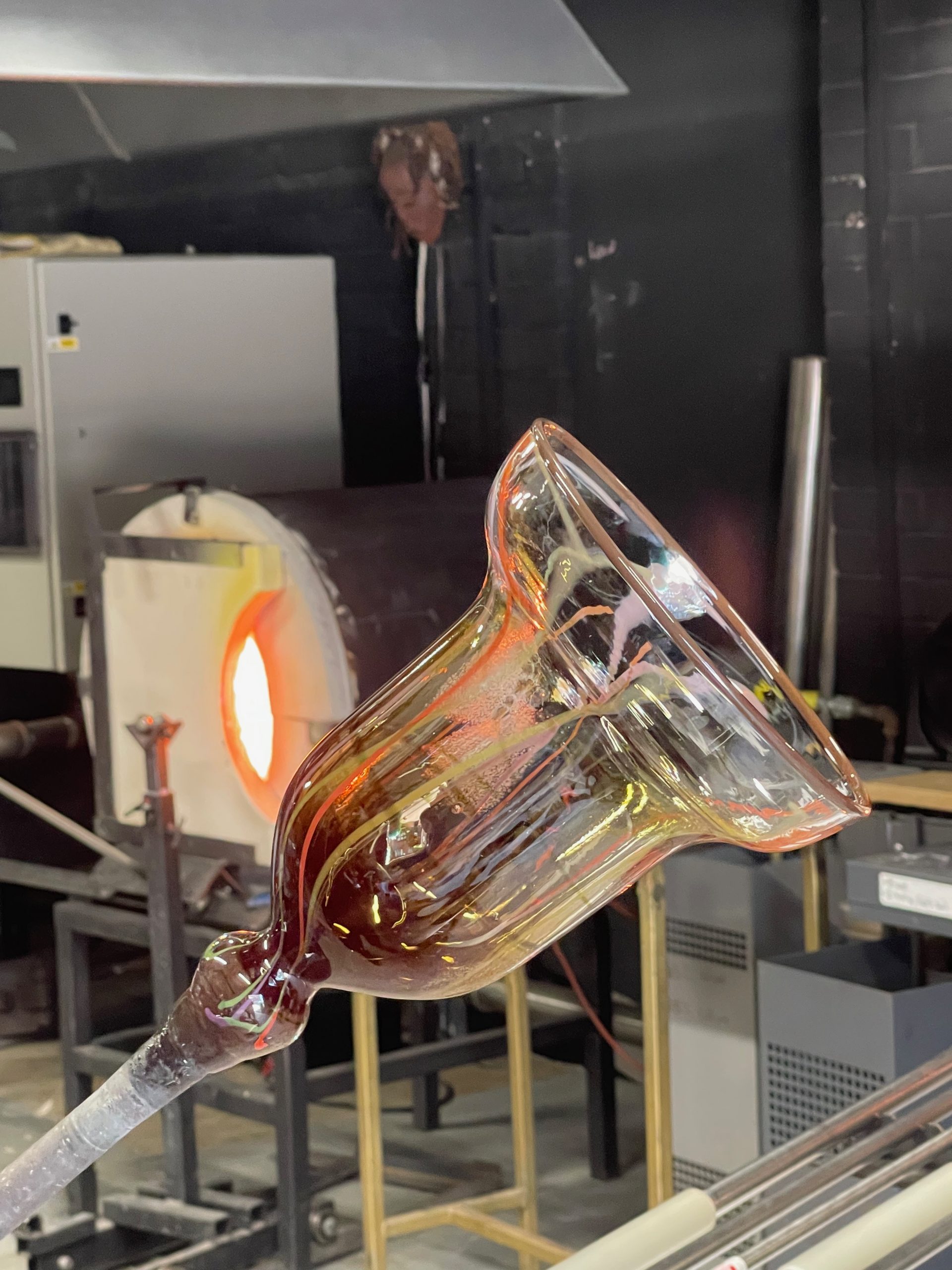 Close-up of a hand-blown glass vessel being shaped on a blowpipe in a glassmaking studio. The transparent form is illuminated by the glow of a furnace in the background.