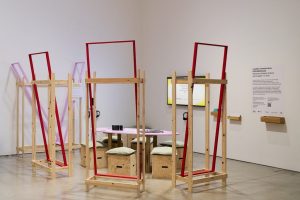 An interactive gallery installation features wooden frames, seating, headphones and a central table arranged within an exhibition space. Wall-mounted displays and interpretive panels surround the installation, inviting visitors to explore digital and physical content.