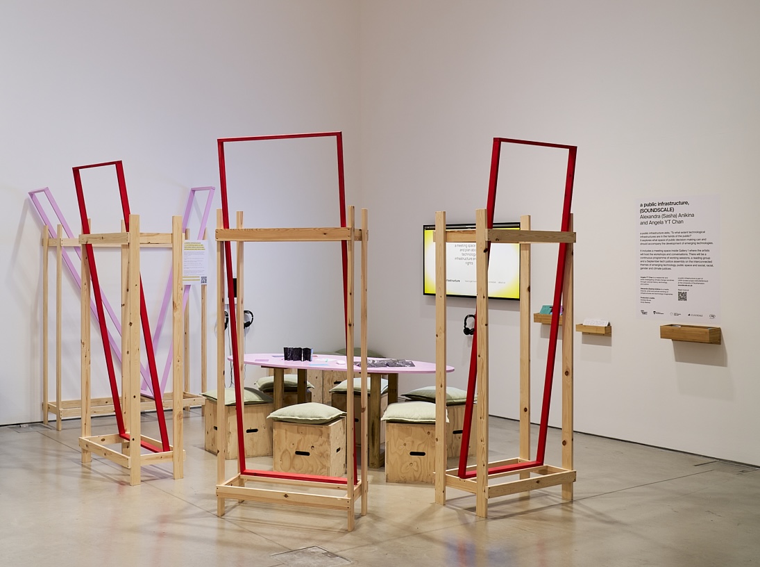 An interactive gallery installation features wooden frames, seating, headphones and a central table arranged within an exhibition space. Wall-mounted displays and interpretive panels surround the installation, inviting visitors to explore digital and physical content.