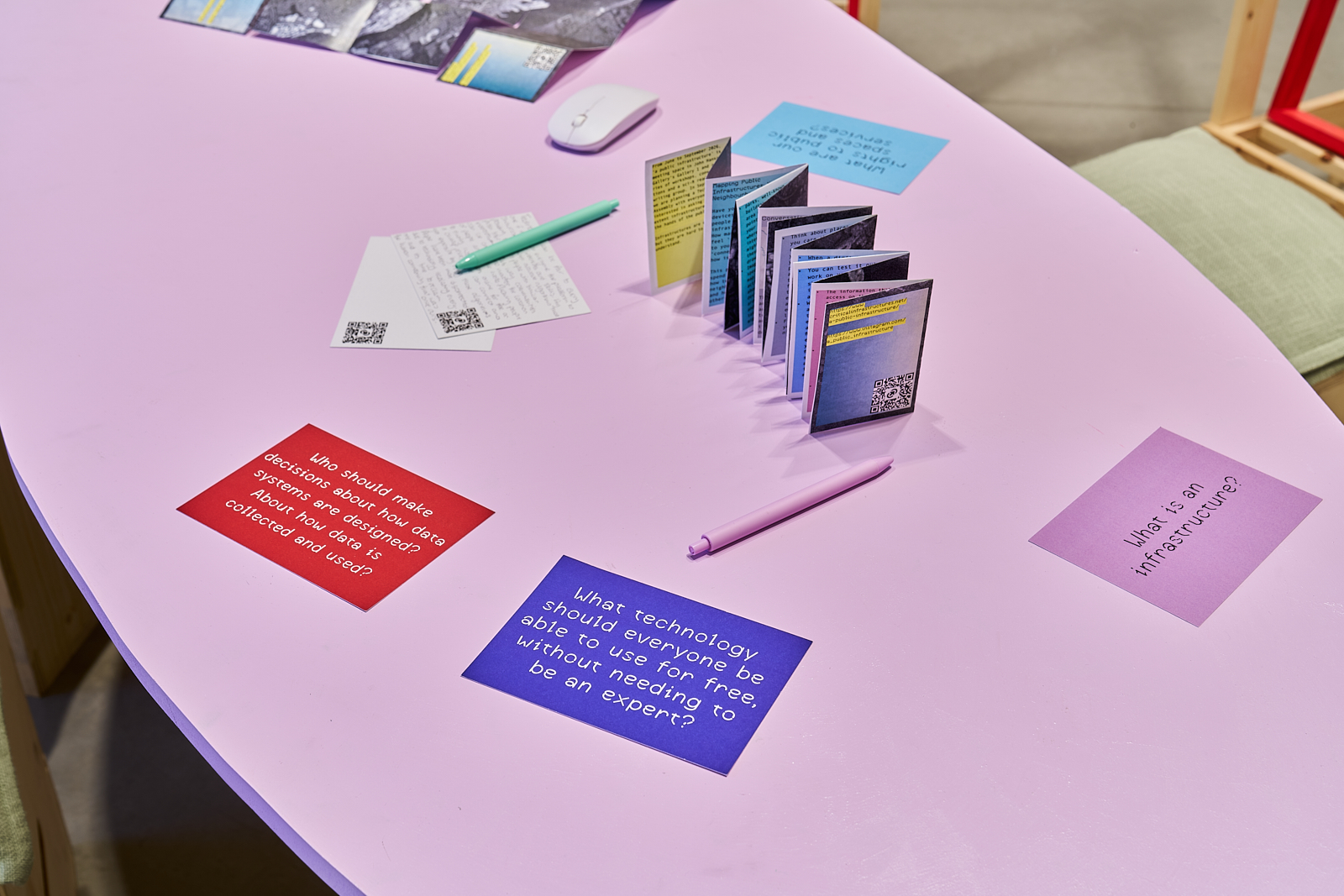 A pink table displays discussion cards, pens, folded booklets and prompts relating to technology, data and infrastructure. The materials are arranged as part of a participatory exhibition activity.