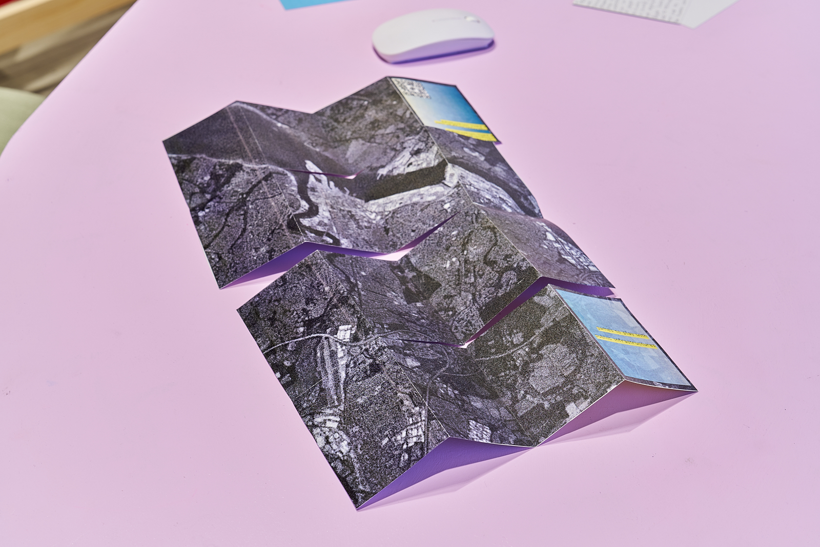 A folded printed leaflet with aerial imagery rests on a pink table beside exhibition materials. The publication appears to be designed for visitors to handle and explore as part of the display.