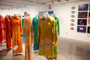 A gallery exhibition displays brightly coloured patterned dresses suspended on mannequins, with photographic artworks and graphic displays mounted on the surrounding walls. The garments feature bold designs in shades of yellow, green, orange, blue and pink.