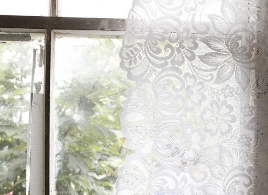 Sunlight filters through a lace curtain hanging in an old window, with foliage visible outside. The image highlights the intricate floral pattern of the lace against the bright light.