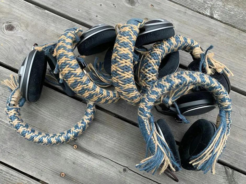 Headphones are wrapped in a long braided blue and beige textile cord, with several loops and tasselled ends arranged on a wooden deck. The artwork combines audio equipment and fibre craft to create a sculptural installation.