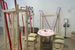 An exhibition installation combines wooden frames, cushioned seating, headphones and a pink table within a gallery space. The arrangement creates a dedicated area for visitors to engage with audio, reading and discussion materials.