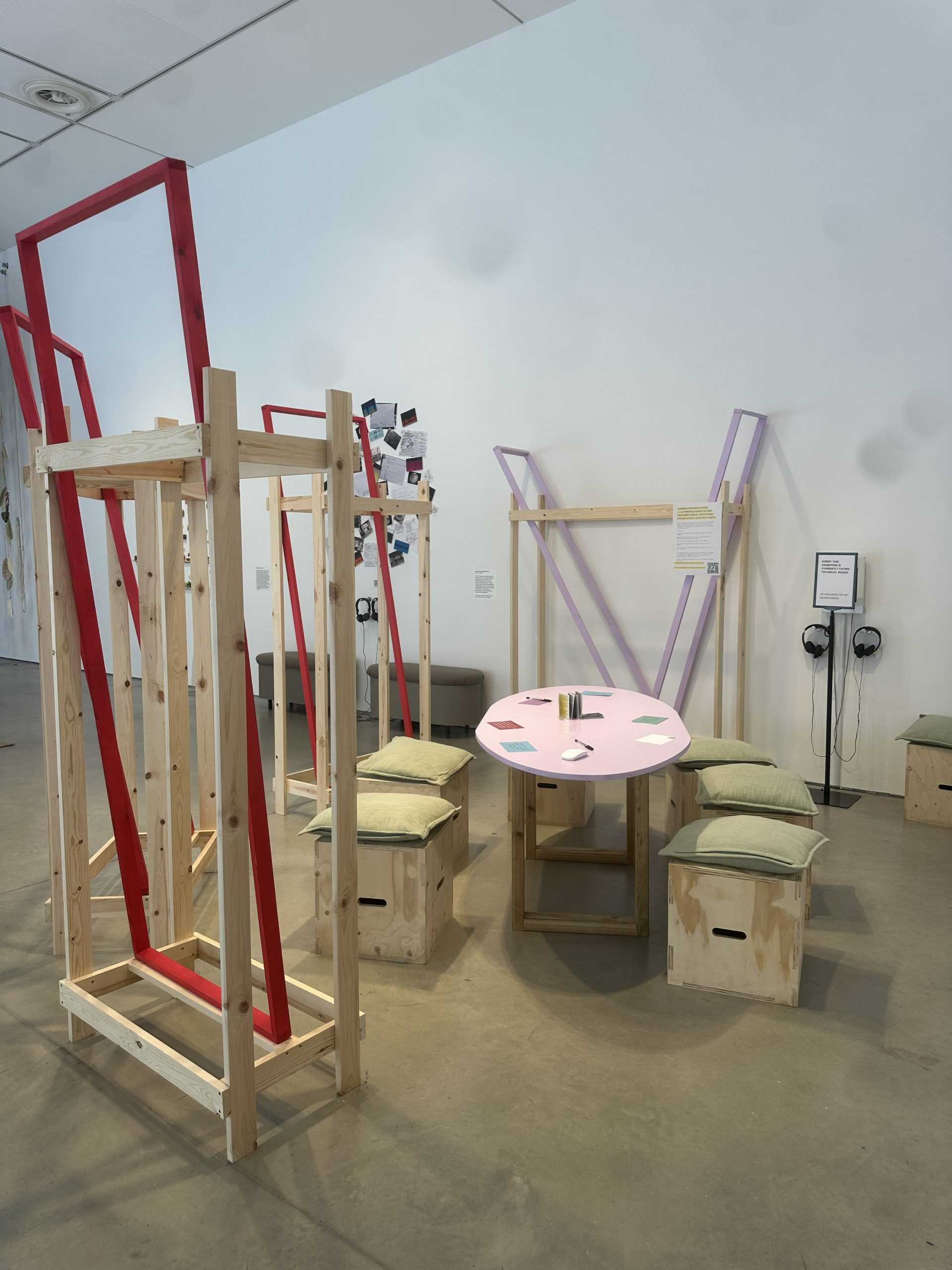 An exhibition installation combines wooden frames, cushioned seating, headphones and a pink table within a gallery space. The arrangement creates a dedicated area for visitors to engage with audio, reading and discussion materials.