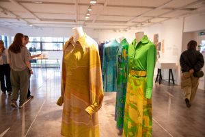 Visitors walk among suspended garments in green, yellow and blue patterned fabrics displayed in a gallery. The dresses are presented on hanging mannequins with artworks visible in the background.