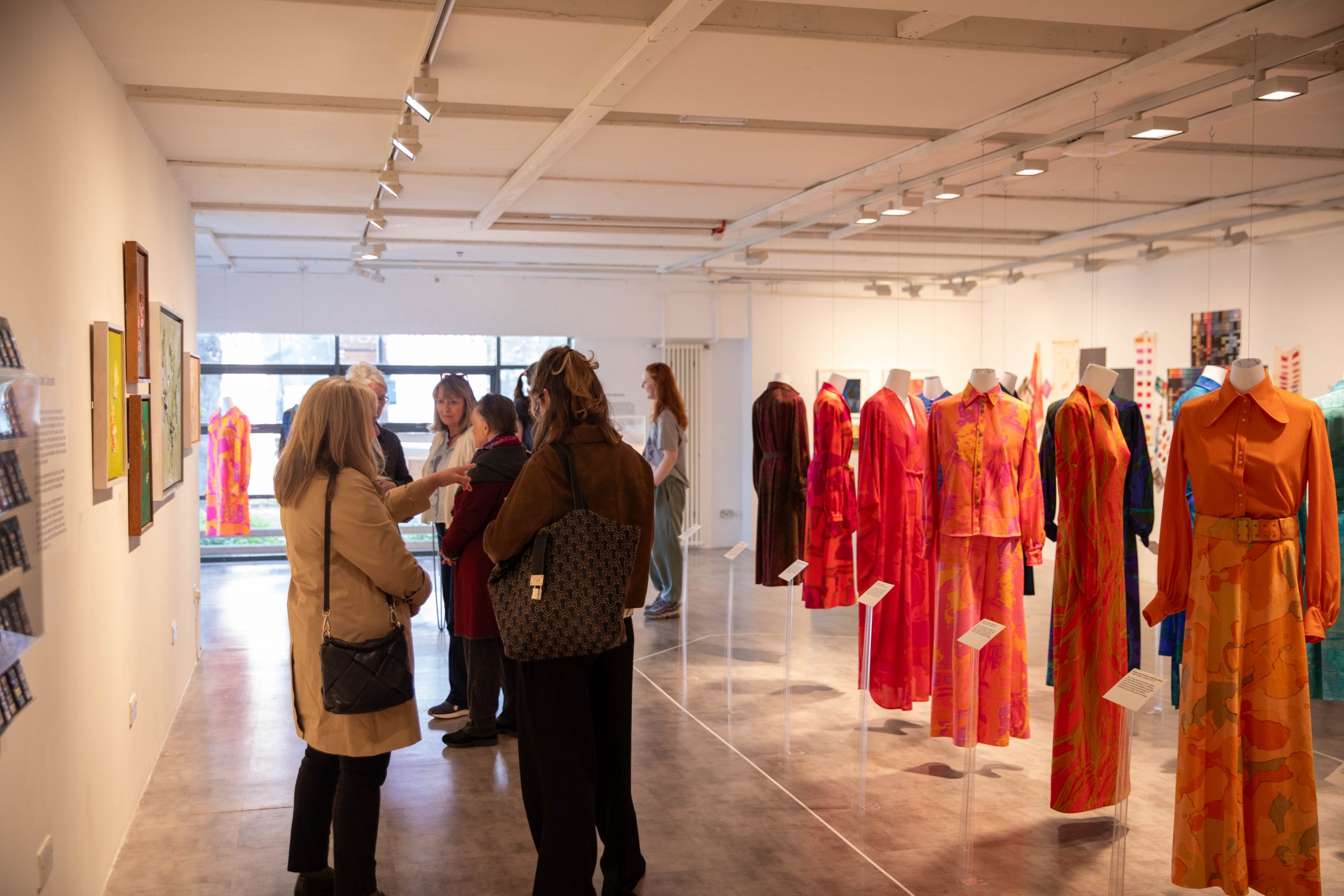 Visitors explore a fashion exhibition featuring brightly coloured dresses suspended throughout a gallery space. Informational labels and framed artworks accompany the display.