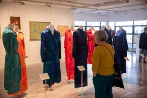 A visitor looks at a collection of colourful patterned dresses displayed on suspended mannequins in a gallery. The exhibition combines fashion design with artworks mounted on the surrounding walls.