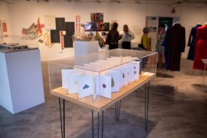 A display case containing an artist’s book or folded publication is shown within a gallery exhibition. Fashion pieces, textile works and visitors are visible in the background.